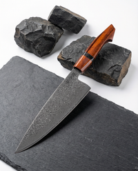 Couteau japonais santoku en acier damas, manche en bois rouge sculpté, posé sur ardoise avec pierres volcaniques.