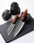 Set de 2 couteaux japonais damas, chef et santoku, manches bois rouge, présentés sur ardoise avec pierres brutes.