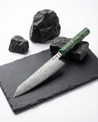 Couteau japonais kiritsuke en acier damas à motif circulaire, manche vert marbré, posé sur ardoise noire avec roches.