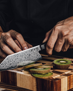 Couteau japonais en acier damassé coupant des tranches de kiwi sur planche en bois à motif damier, tenu en main sur fond sombre.