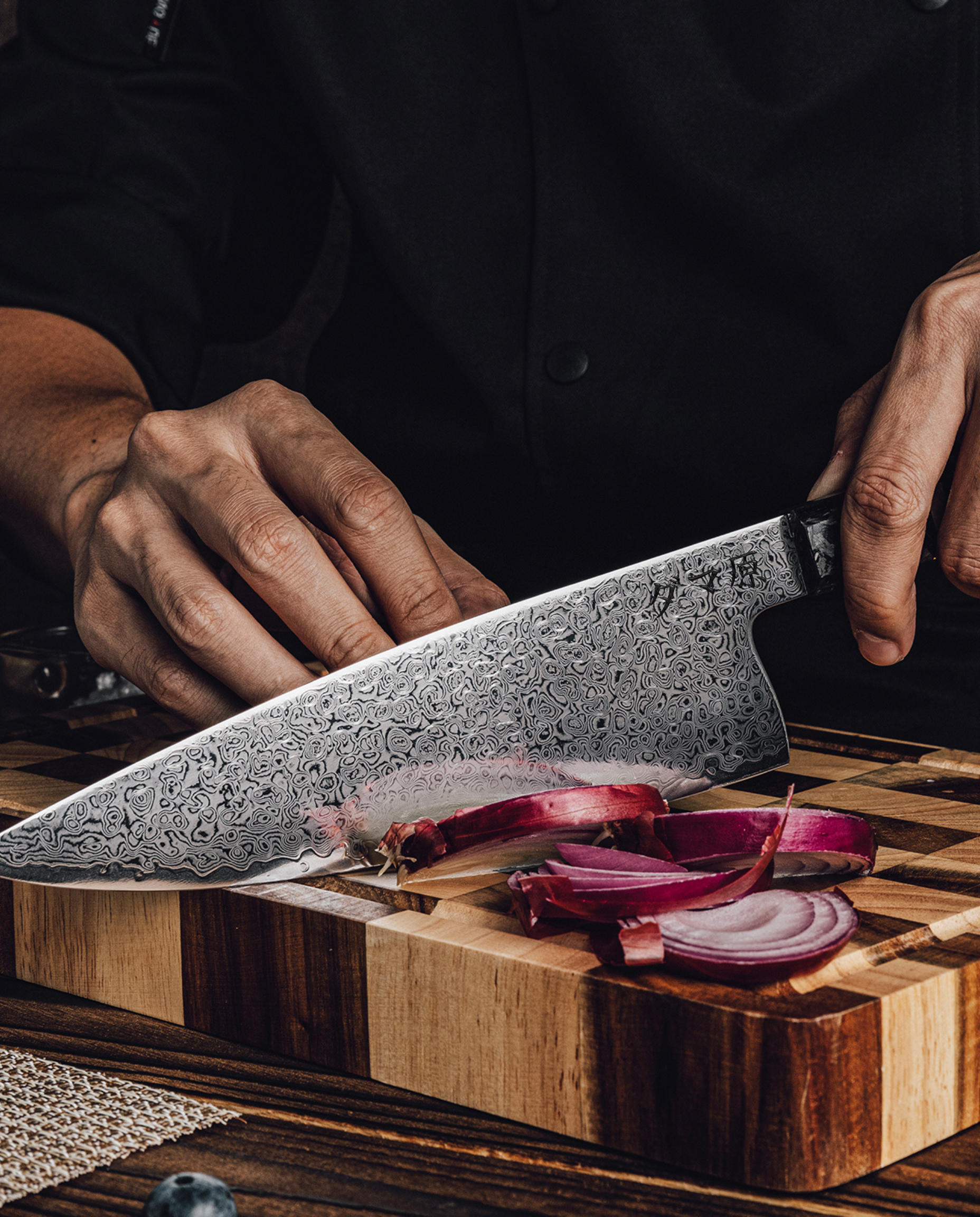 Couteau japonais en acier damassé tranchant un oignon rouge sur planche en bois épaisse, tenu en main sur fond sombre.