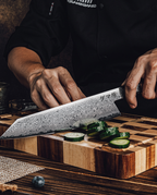 Couteau japonais en acier damassé coupant des rondelles de concombre sur planche en bois massif, tenu en main sur fond sombre.