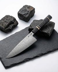 Couteau japonais en acier damassé à manche noir veiné avec bague claire, posé sur ardoise noire entourée de pierres brutes