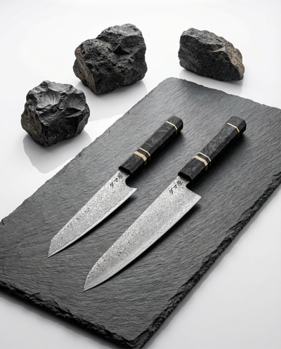 Deux couteaux japonais en acier damassé avec manches noirs veinés, posés sur ardoise noire entourée de pierres brutes