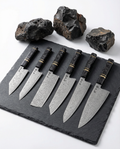 Set complet de 6 couteaux japonais en acier damas, manches noirs texturés avec anneaux dorés, présentés sur ardoise noire.
