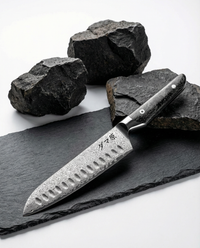 Couteau japonais santoku en acier damassé avec alvéoles, manche noir riveté, posé sur ardoise sombre et pierres brutes