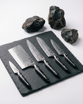 Set de 5 couteaux japonais en acier damassé avec manches noirs, disposés sur ardoise sombre entourée de pierres brutes