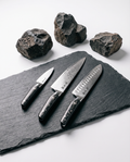 Set de trois couteaux japonais en acier damassé, manches noirs rivetés, posés sur ardoise avec pierres brutes
