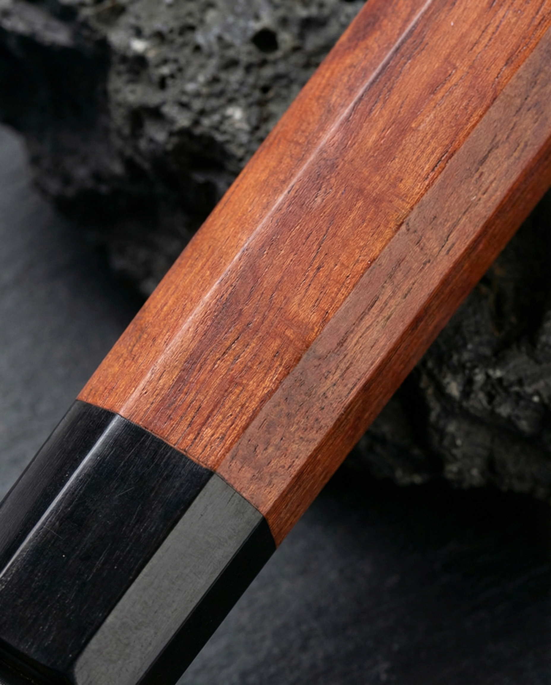 Gros plan sur manche de couteau japonais en bois brun à veinage fin, avec embout noir, posé sur ardoise et pierre sombre
