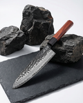 Couteau japonais en acier martelé, manche en bois brun avec virole noire, posé sur ardoise noire entouré de pierres brutes
