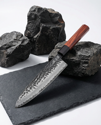 Couteau japonais en acier martelé, manche en bois brun avec virole noire, posé sur ardoise noire entouré de pierres brutes