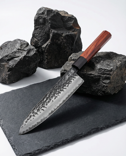Couteau japonais en acier martelé, manche en bois brun avec virole noire, posé sur ardoise noire entouré de pierres brutes