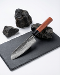 Couteau japonais en acier martelé, manche en bois brun avec virole noire, posé sur ardoise noire avec pierres volcaniques