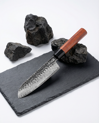 Couteau japonais en acier martelé, manche en bois brun avec virole noire, posé sur ardoise noire avec pierres volcaniques