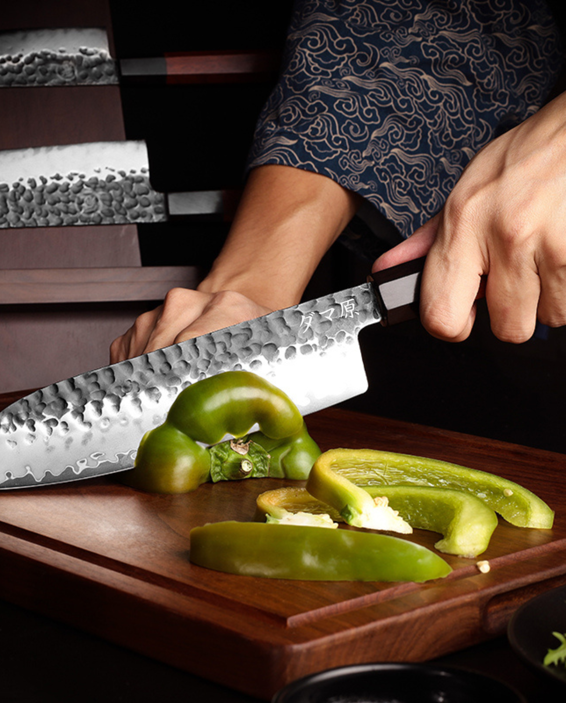Couteau japonais en acier damassé martelé découpant un poivron vert sur planche en bois, main tenant le manche, support à couteaux en arrière-plan
