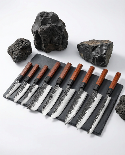Set de neuf couteaux japonais en acier martelé, manches en bois foncé, alignés sur ardoise avec pierres brutes