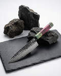 Couteau japonais Damas Origin avec lame en acier Damas 67 couches finition martelée et gravure japonaise, manche en bois stabilisé vert et pourpre avec insert métallique, posé sur plaque d’ardoise noire entourée de pierres volcaniques.
