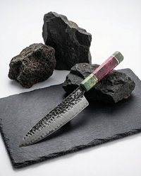 Couteau japonais Damas Origin avec lame en acier Damas 67 couches finition martelée et gravure japonaise, manche en bois stabilisé vert et pourpre avec insert métallique, posé sur plaque d’ardoise noire entourée de pierres volcaniques.