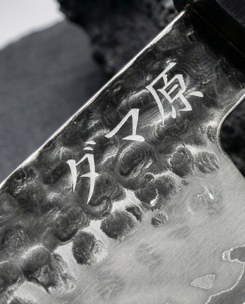 Couteau japonais en acier martelé, lame gravée de caractères japonais, posé sur ardoise sombre avec pierre en arrière-plan

