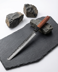Couteau japonais en acier martelé à lame longue, manche en bois brun, posé sur ardoise noire avec pierres brutes