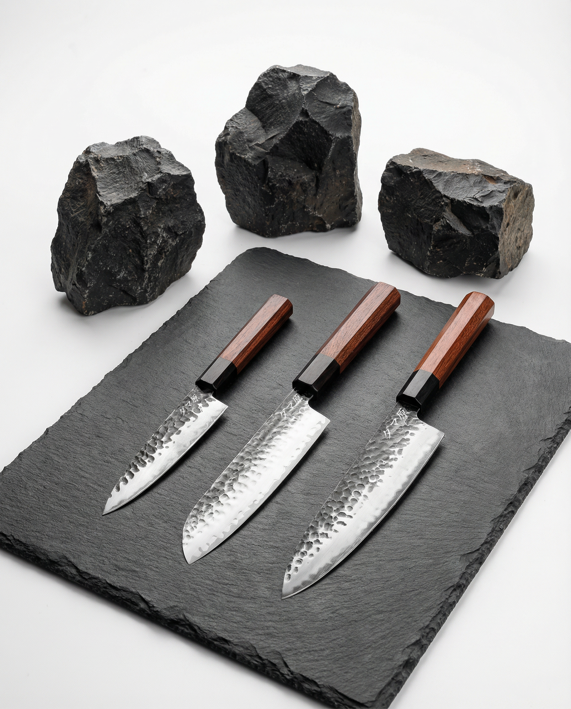 Set de trois couteaux japonais en acier martelé, manches en bois brun, posés sur ardoise avec pierres brutes