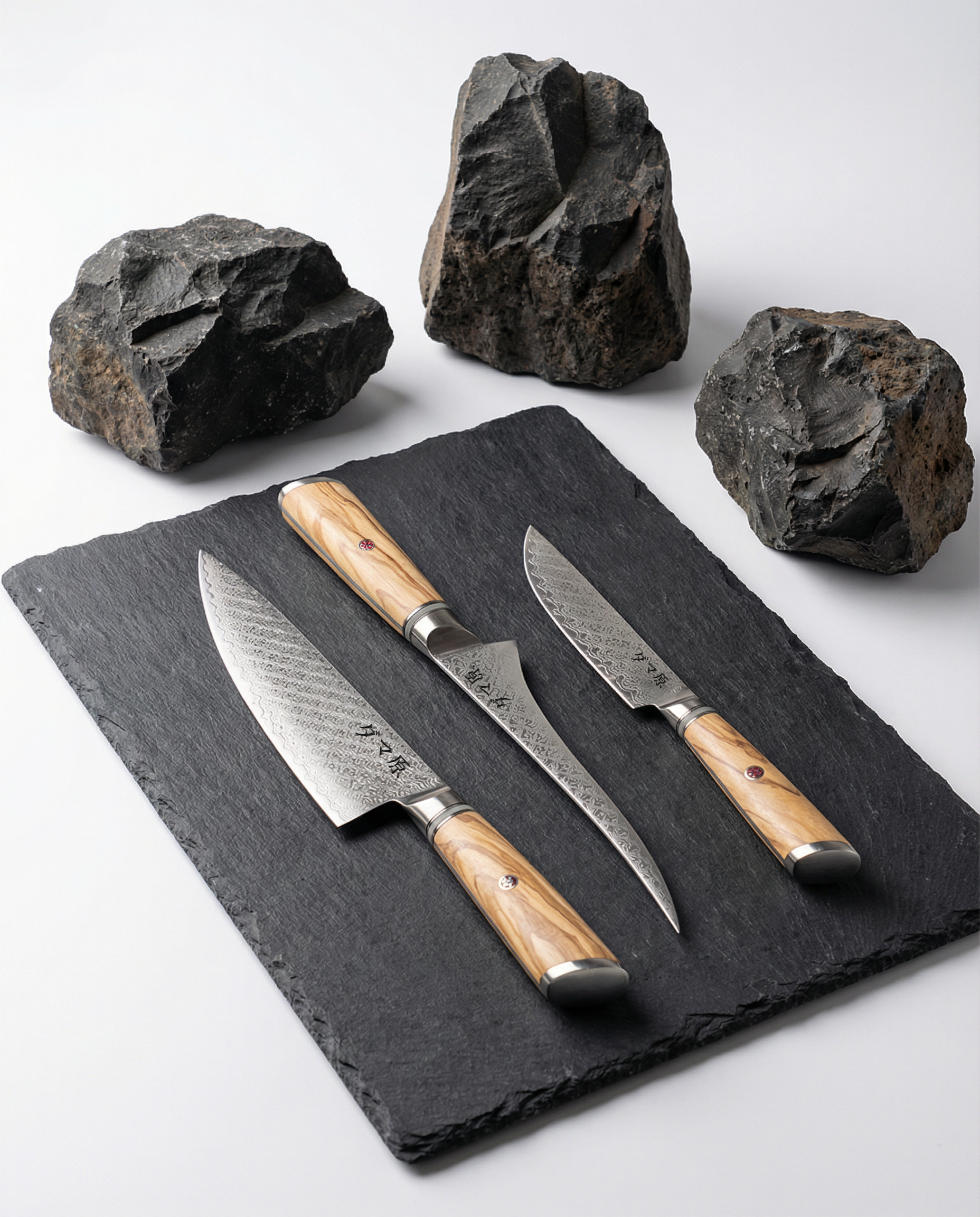 Set de 3 couteaux japonais en acier damas, manches bois clair veinés, présentés sur plaque d’ardoise noire avec roches brutes.