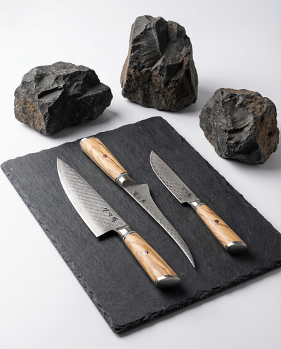 Set de 3 couteaux japonais en acier damas, manches bois clair veinés, présentés sur plaque d’ardoise noire avec roches brutes.